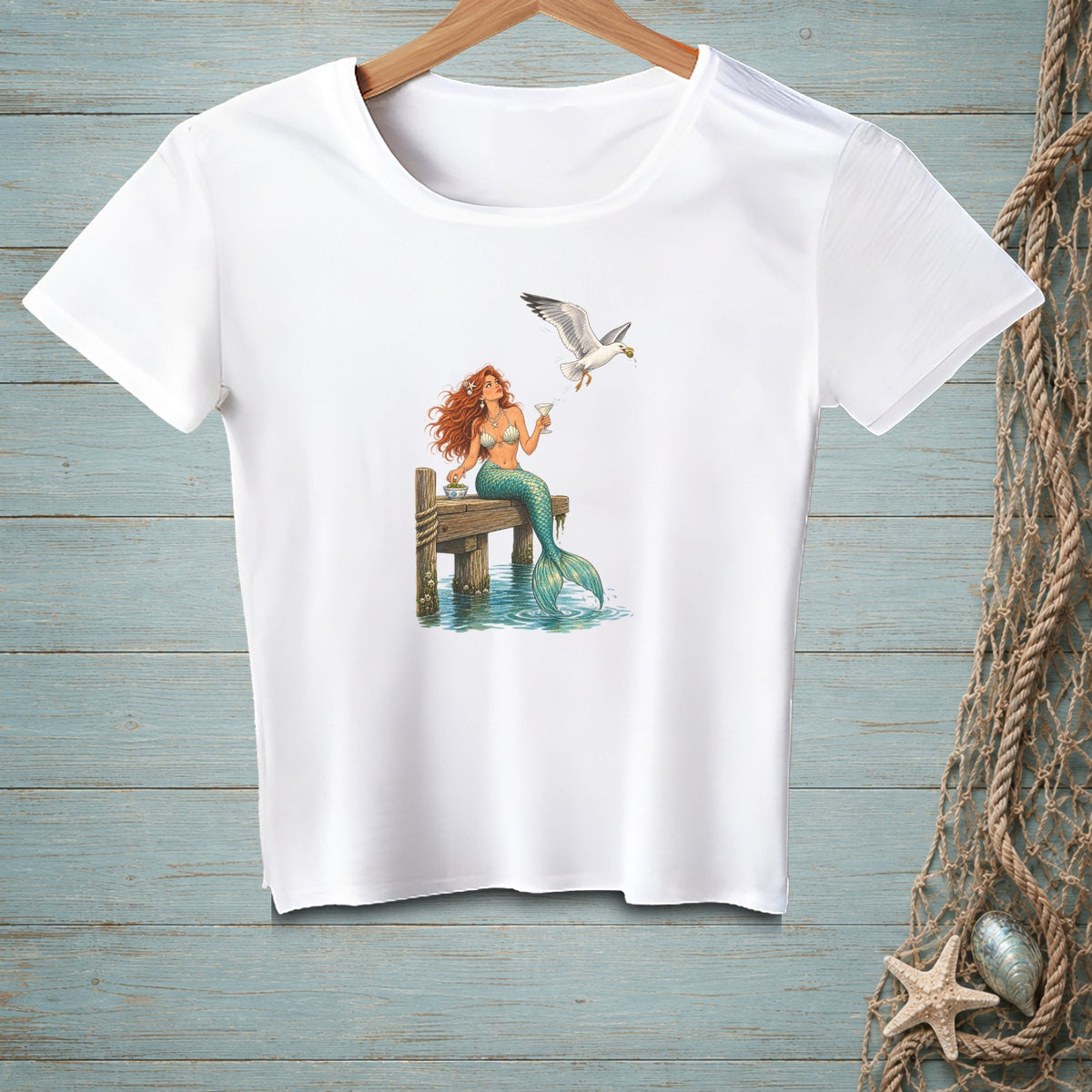 White t-shirt with a mermaid and seagull graphic on a wooden dock.