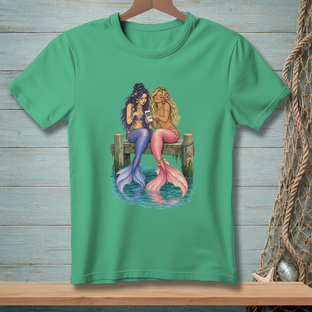 Two mermaids with colorful tails sit on a wooden dock looking at a smartphone.