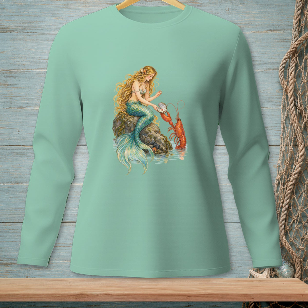 Mermaid sitting on a rock with a lobster and a pearl on a light green long sleeve shirt.