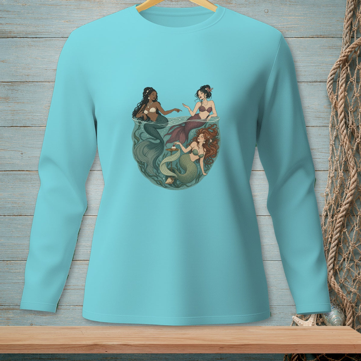 Three mermaids swimming in a circle with seaweed and shells in a teal long-sleeve shirt.