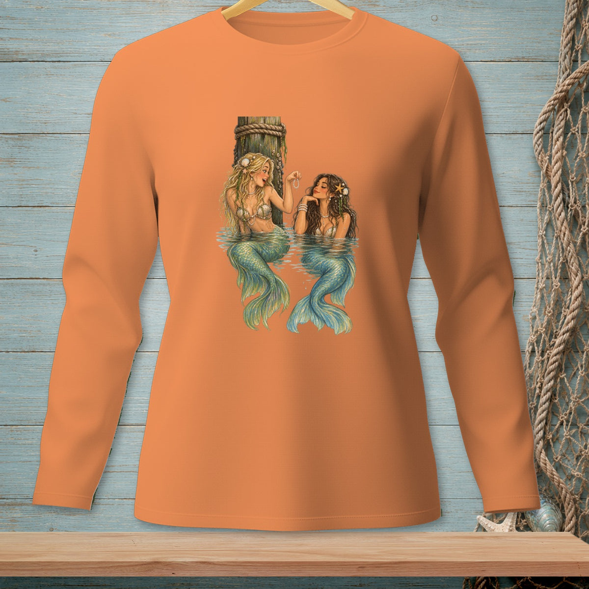 Two mermaids with blue and green tails sit near a wooden post by the water.