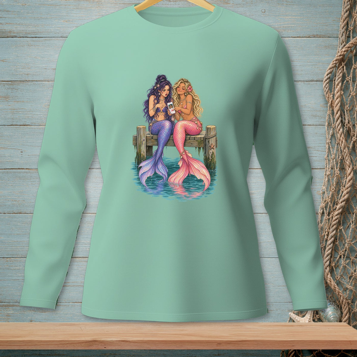 Two mermaids with colorful tails sit on a dock, looking at a smartphone.