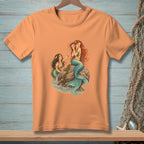 Two mermaids with blue and green tails sit on rocks by the ocean.