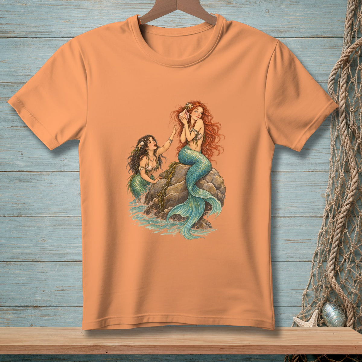 Two mermaids with blue and green tails sit on rocks by the ocean.