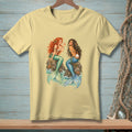 Pale yellow t-shirt with two illustrated mermaids sitting on rocks, holding a pearl.