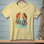 Pale yellow t-shirt with two illustrated mermaids sitting on rocks, holding a pearl.