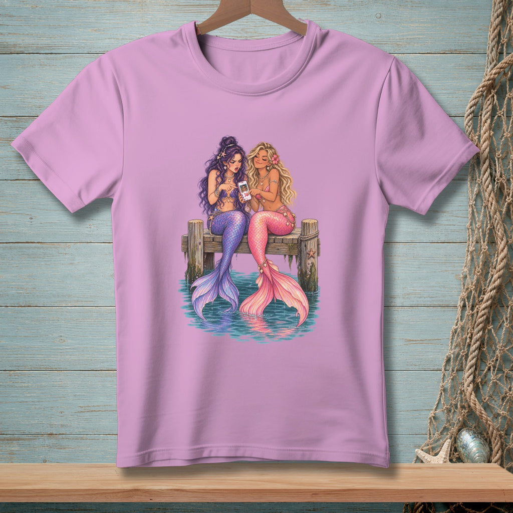 Two mermaids with purple and pink tails sit on a dock looking at a smartphone.