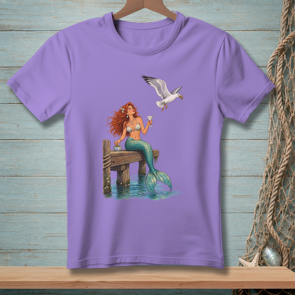 Lavender t-shirt with a mermaid holding a drink near a seagull on a dock.